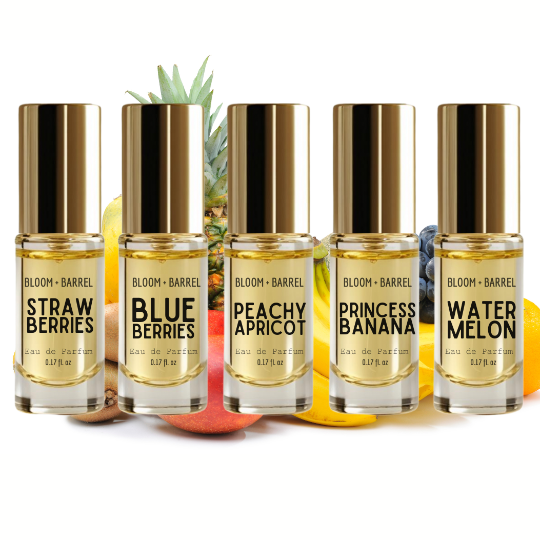 Six fragrance oil bottles from BLOOM + BARREL lined up on a white background with fruit props behind them. The bottles are labeled Strawberry, Blueberries, Peachy Apricot, Princess Banana, and Watermelon, and feature gold caps and clear glass showing a golden-yellow liquid inside.