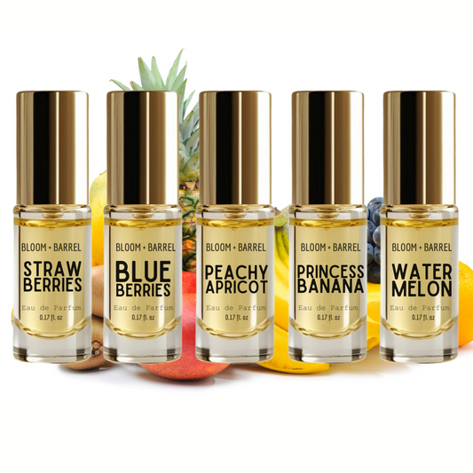 Six fragrance oil bottles from BLOOM + BARREL lined up on a white background with fruit props behind them. The bottles are labeled Strawberry, Blueberries, Peachy Apricot, Princess Banana, and Watermelon, and feature gold caps and clear glass showing a golden-yellow liquid inside.