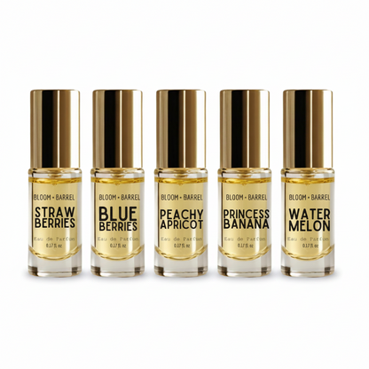 Five small glass perfume bottles with shiny gold caps arranged in a row on a white background. The labels read BLOOM + BARREL and the scents: Strawberries, Blueberries, Peachy Apricot, Princess Banana, and Watermelon.