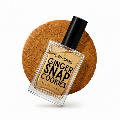 A square glass perfume bottle labeled 'BLOOM • BARREL GINGER SNAP COOKIES Eau de Parfum' with a black cap, centered in front of a round gingerbread cookie on a white background.