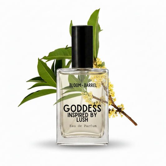 A rectangular glass perfume bottle labeled 'GODDESS INSPIRED BY LUSH' and 'BLOOM • BARREL' with a black cap, shown on a white background with green leaves and small yellow flowers behind it.