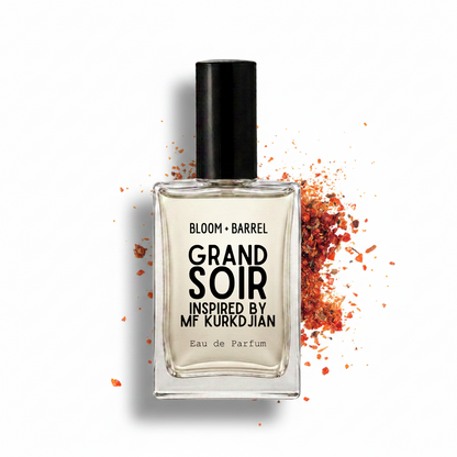 A clear rectangular perfume bottle with a black cap and black text reading 'BLOOM • BARREL GRAND SOIR INSPIRED BY MF KURDJIAN Eau de Parfum,' centered on a white background with scattered orange-red spice-like flakes around it.