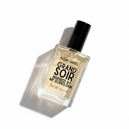 A rectangular glass perfume bottle with a black cap, containing a pale yellow liquid. The front label reads 'BLOOM • BARREL GRAND SOIR INSPIRED BY MF KURKDJIAN Eau de Parfum' in black text, set against a plain white background.