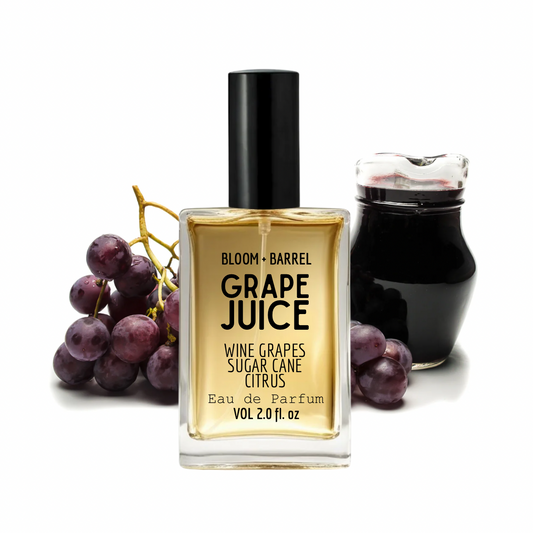 Bloom + Barrel Grape Juice Eau de Parfum bottle in the center, flanked by a bunch of purple grapes on the left and a dark glass jar on the right, all on a white background.