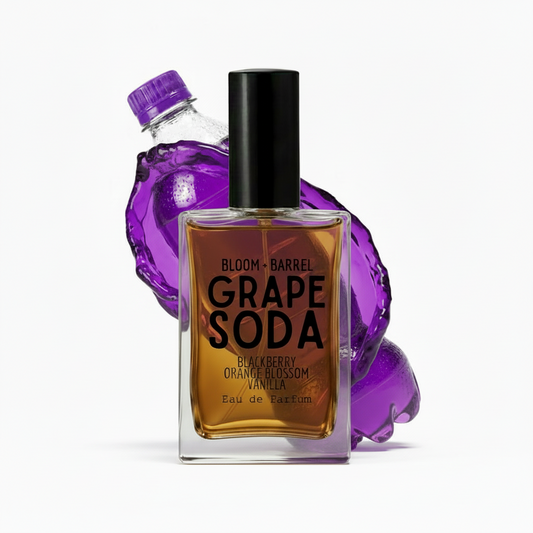 A bottle of Bloom + Barrel Grape Soda Eau de Parfum with a black cap, set against a white background with a purple grape soda splash and a clear bottle behind it.