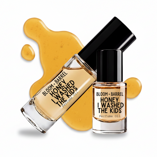 Two glass bottles of Bloom + Barrel 'Honey I Washed The Kids' perfume oil with black caps, arranged over a glossy amber honey smear on a white background.