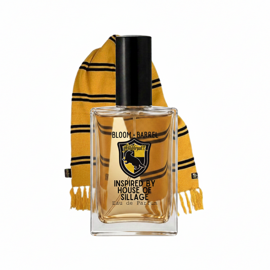 A rectangular glass perfume bottle with a black cap and amber liquid, labeled 'BLOOM + BARREL' and 'INSPIRED BY HOUSE OF SILLAGE', shown in front of a yellow and black striped scarf on a white background.