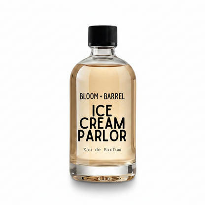 A clear glass perfume bottle with a black cap, filled with a pale amber liquid. The bottle features a white label with black text reading 'BLOOM + BARREL' and 'ICE CREAM PARLOR Eau de Parfum'.