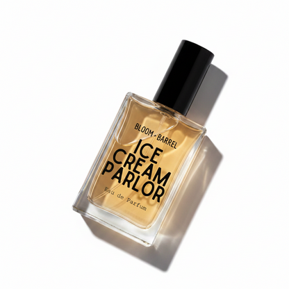A rectangular glass perfume bottle with a black cap, filled with amber-colored fragrance liquid. The front label reads 'BLOOM • BARREL ICE CREAM PARLOR Eau de Parfum' in black text, set against a plain white background.