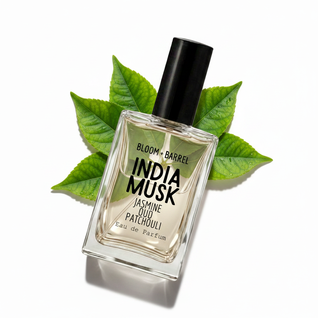 A rectangular glass perfume bottle labeled 'BLOOM + BARREL INDIA MUSK Jasmine Oud Patchouli Eau de Parfum' with a black cap, centered on a white background and surrounded by scattered green leaves.