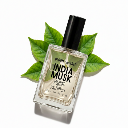 A rectangular glass perfume bottle labeled 'BLOOM + BARREL INDIA MUSK Jasmine Oud Patchouli Eau de Parfum' with a black cap, centered on a white background and surrounded by scattered green leaves.