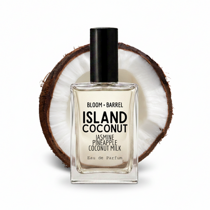 A rectangular glass perfume bottle labeled 'BLOOM • BARREL ISLAND COCONUT' with a black cap, centered in front of a halved coconut on a white background.