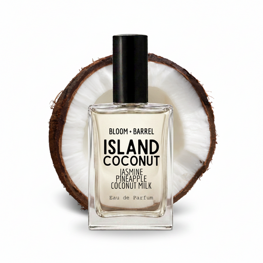 A rectangular glass perfume bottle labeled 'BLOOM • BARREL ISLAND COCONUT' with a black cap, centered in front of a halved coconut on a white background.
