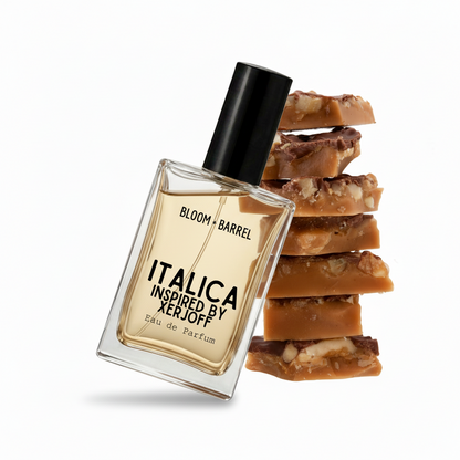 A clear rectangular perfume bottle with a black cap and amber-gold liquid, labeled 'ITALICA INSPIRED BY XERJOFF' and 'BLOOM • BARREL', shown in front of stacked caramel and cookie pieces on a white background.