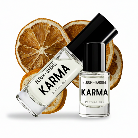 Two clear glass bottles of Bloom + Barrel Karma Perfume Oil with black caps, arranged in front of dried orange slices on a white background.