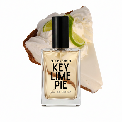 A rectangular glass perfume bottle labeled 'BLOOM + BARREL KEY LIME PIE Eau de Parfum' with a black cap, centered in front of a dessert slice with whipped cream, lime, and graham cracker on a white background.