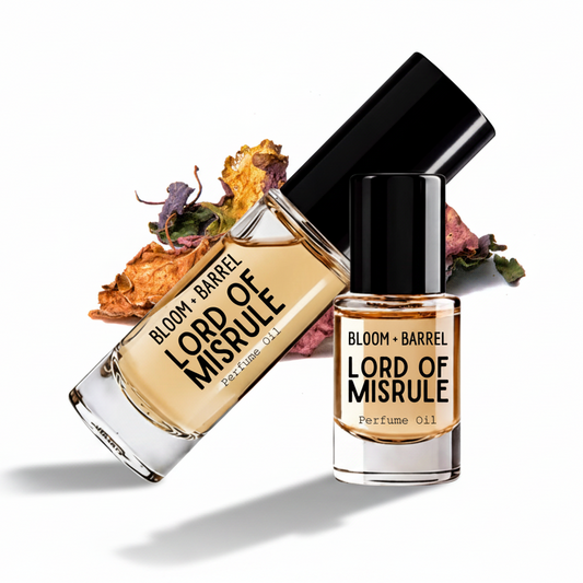 Two glass bottles of BLOOM + BARREL Lord of Misrule Perfume Oil with black caps, one upright and one tilted, with dried rose petals in the background on a white studio background.