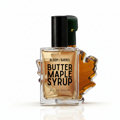 Bloom Barrel Butter Maple Syrup Eau de Parfum bottle with a clear glass body showing amber liquid, a black cap, and a maple leaf-shaped accent on a white background.