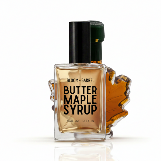 Bloom Barrel Butter Maple Syrup Eau de Parfum bottle with a clear glass body showing amber liquid, a black cap, and a maple leaf-shaped accent on a white background.
