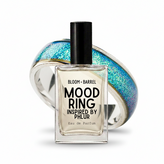 A clear glass perfume bottle labeled 'BLOOM • BARREL MOOD RING INSPIRED BY PHLR Eau de Parfum' with a black cap, set in front of a translucent ring featuring blue, green, and gold glittery segments on a white background.