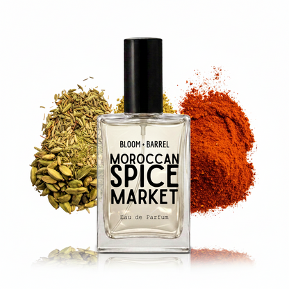 A clear glass perfume bottle labeled 'BLOOM • BARREL MOROCCAN SPICE MARKET Eau de Parfum' with a black cap, centered on a white background and flanked by piles of green cardamom and red chili powder.