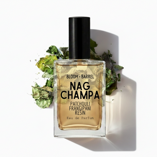 A rectangular glass bottle of Nag Champa patchouli frangipani resin perfume with a black cap, centered on a white background with dried green and yellow botanicals behind it. Visible text on the bottle reads 'BLOOM • BARREL' and 'NAG CHAMPA'.