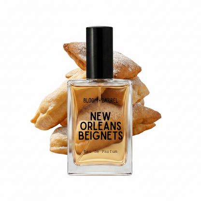 A rectangular glass perfume bottle with a black cap and amber liquid, labeled 'BLOOM • BARREL NEW ORLEANS BEIGNETS Eau de Parfum', centered in front of a pile of powdered pastries on a white background.