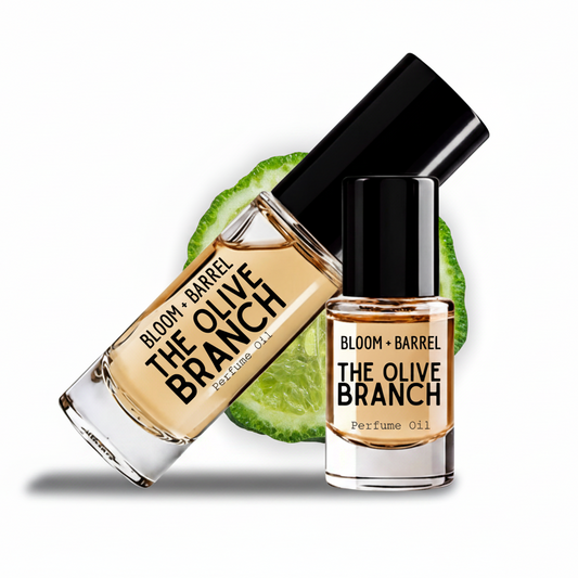 Two glass bottles of Bloom + Barrel The Olive Branch perfume oil with black caps, one larger bottle tilted diagonally and one smaller upright bottle, both showing the amber-colored oil inside. A sliced green olive is placed behind the bottles on a clean white background.