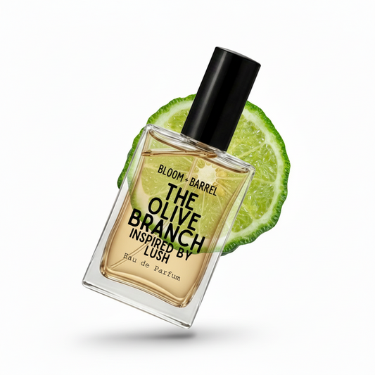 A rectangular glass perfume bottle with a black cap, filled with amber-toned fragrance, labeled 'THE OLIVE BRANCH' by Bloom + Barrel. A sliced lime is placed behind the bottle on a clean white background.