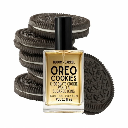 A bottle of Bloom + Barrel Oreo Cookies Eau de Parfum in a square glass bottle with a black cap, set against a background of stacked and leaning Oreo cookies. The liquid inside the bottle is a warm golden-amber color.