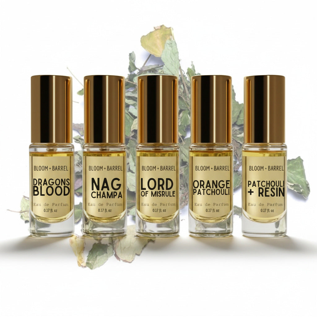Five Bloom + Barrel perfume bottles arranged in a row on a white background with dried botanical leaves behind them. The bottles contain pale golden liquid and have shiny metallic gold caps.