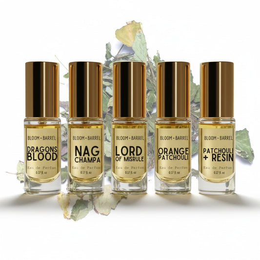 Five Bloom + Barrel perfume bottles arranged in a row on a white background with dried botanical leaves behind them. The bottles contain pale golden liquid and have shiny metallic gold caps.