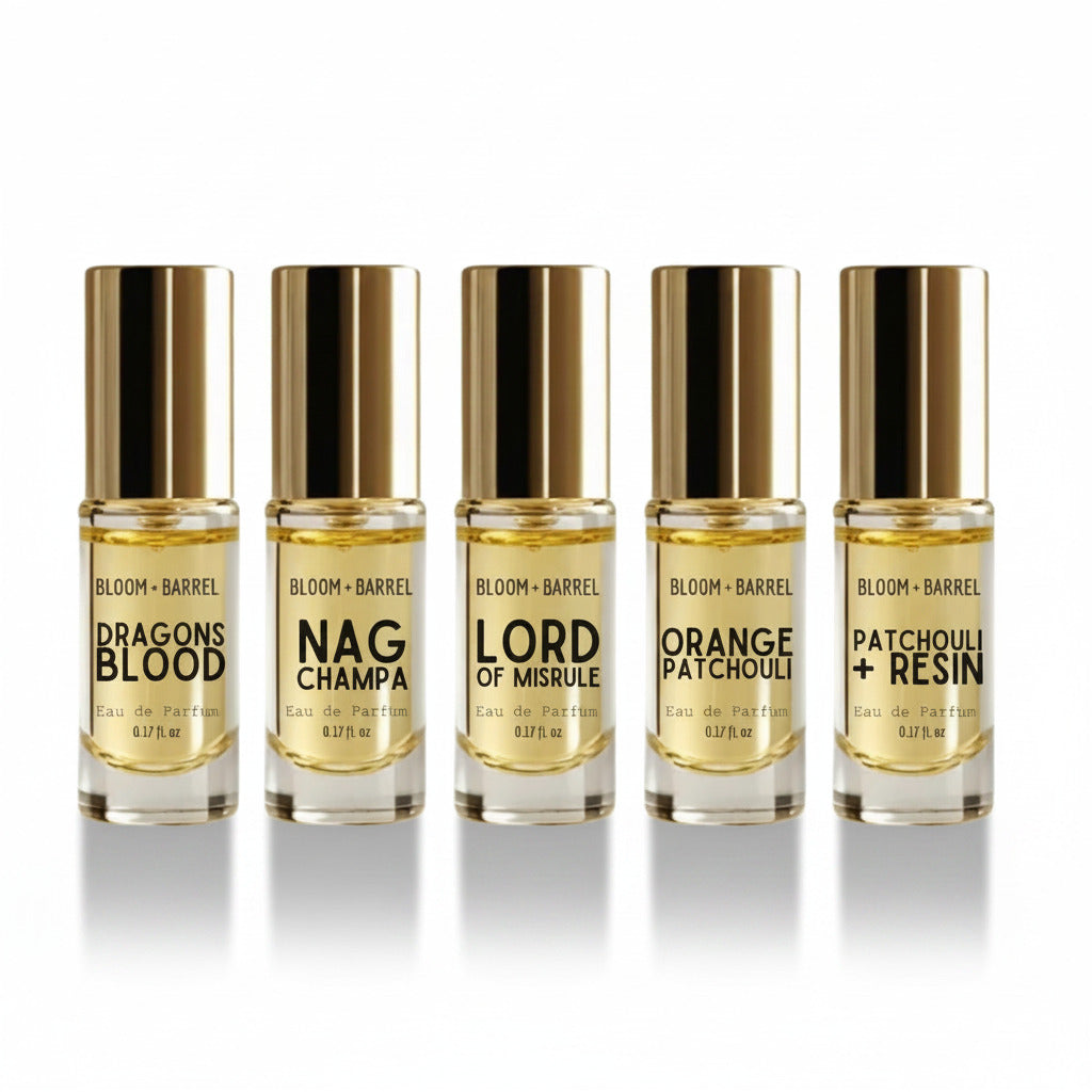 Five small perfume bottles with gold caps arranged in a row on a white background. The labels read BLOOM + BARREL and the scents Dragons Blood, Nag Champa, Lord of Misrule, Orange Patchouli, and Patchouli + Resin.