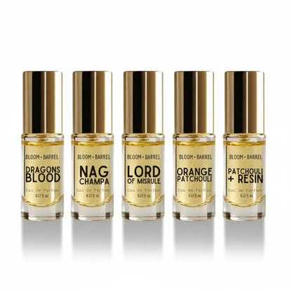 Five small perfume bottles with gold caps arranged in a row on a white background. The labels read BLOOM + BARREL and the scents Dragons Blood, Nag Champa, Lord of Misrule, Orange Patchouli, and Patchouli + Resin.