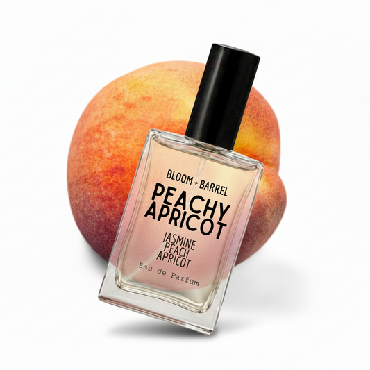 A rectangular glass perfume bottle labeled 'BLOOM + BARREL PEACHY APRICOT' stands in front of two whole apricots on a clean white background.