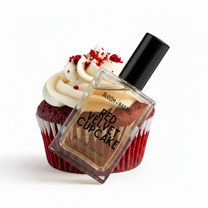 A perfume bottle labeled 'BLOOM • BARREL RED VELVET CUPCAKE Eau de Parfum' centered in front of a red velvet cupcake with white frosting and red crumbs, on a plain white background.
