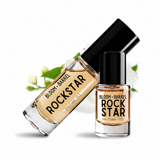 Two glass bottles of Bloom + Barrel Rockstar Perfume Oil with black caps, one standing upright and one tilted diagonally, both showing the amber-colored oil inside. White flower buds are placed behind the bottles on a clean white background.