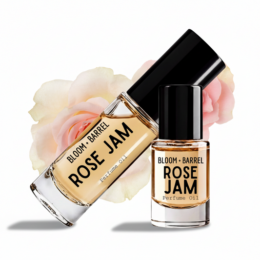Two glass bottles of Bloom + Barrel Rose Jam perfume oil with black caps, one larger bottle lying diagonally and one smaller upright bottle, both filled with amber-toned liquid and set against soft cream and pink rose petals on a white background.