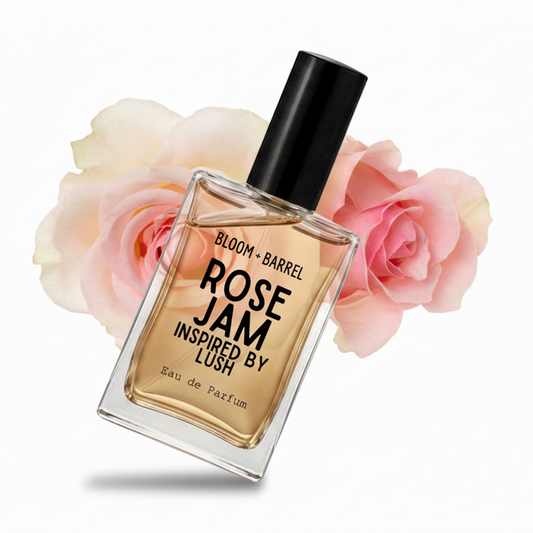 A rectangular glass perfume bottle with a black cap, labeled 'BLOOM • BARREL ROSE JAM INSPIRED BY LUSH', centered in front of soft pink roses on a white background.