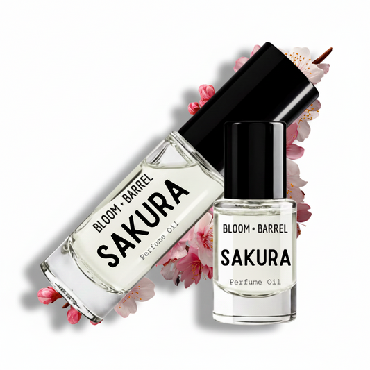 Two clear glass perfume oil bottles with black caps from BLOOM + BARREL, labeled SAKURA, arranged on a white background with pink cherry blossoms behind them.