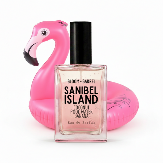 A rectangular glass perfume bottle labeled 'BLOOM • BARREL SANIBEL ISLAND Coconut Pool Water Banana Eau de Parfum' with a black cap, centered in front of a bright pink inflatable flamingo float on a white background.
