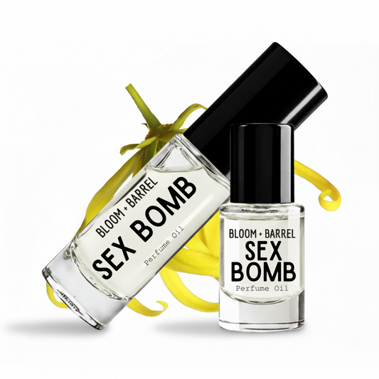 Two clear glass perfume oil bottles with black caps from Bloom + Barrel, labeled 'SEX BOMB Perfume Oil,' arranged on a white background with yellow ribbon accents.