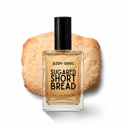 A square glass perfume bottle labeled 'BLOOM • BARREL SUGARED SHORT BREAD Eau de Parfum' with a black cap, set in front of a sugar-dusted shortbread cookie on a white background.