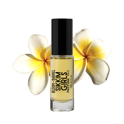 A bottle of Sikkim Girls perfume oil by Bloom + Barrel, featuring a black cap and clear glass bottle showing the pale yellow oil inside, set against a white background with two yellow and white plumeria flowers behind it.