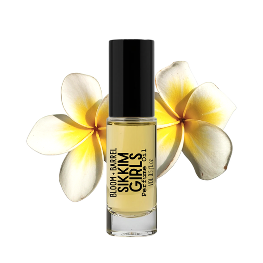 A bottle of Sikkim Girls perfume oil by Bloom + Barrel, featuring a black cap and clear glass bottle showing the pale yellow oil inside, set against a white background with two yellow and white plumeria flowers behind it.