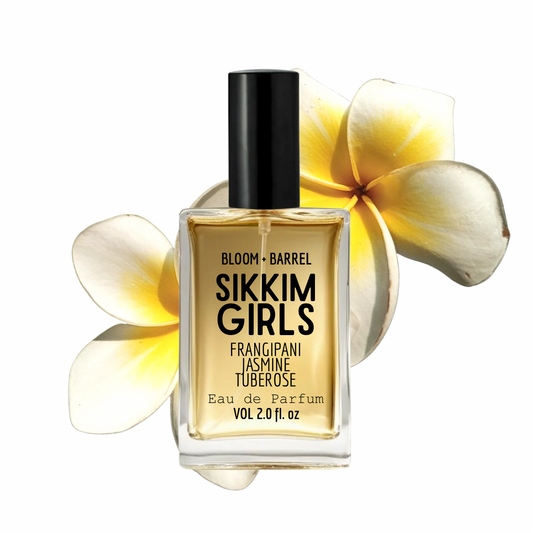 A rectangular glass perfume bottle with a black cap, labeled 'BLOOM + BARREL SIKKIM GIRLS', filled with golden-yellow fragrance. The bottle is centered against a white background with two large pale yellow and cream plumeria flowers behind it.