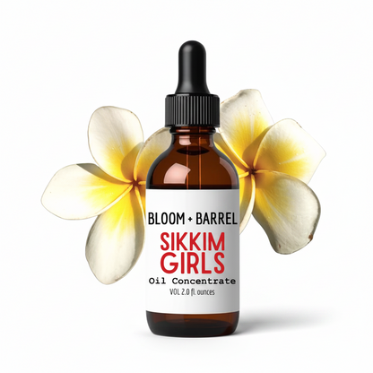 An amber glass dropper bottle labeled BLOOM + BARREL Sikkim Girls Oil Concentrate, centered on a white background with two yellow-and-white plumeria flowers behind it.