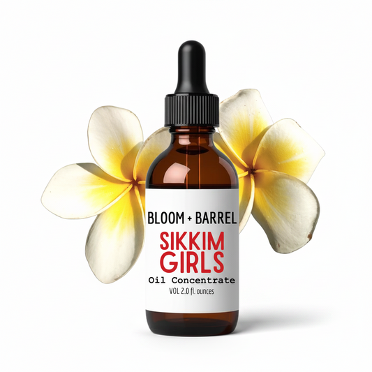 An amber glass dropper bottle labeled BLOOM + BARREL Sikkim Girls Oil Concentrate, centered on a white background with two yellow-and-white plumeria flowers behind it.