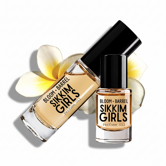 Two glass bottles of Sikkim Girls perfume oil by Bloom + Barrel, one upright and one tilted, with a black cap and amber-colored oil inside, styled with white and yellow plumeria flowers on a white background.