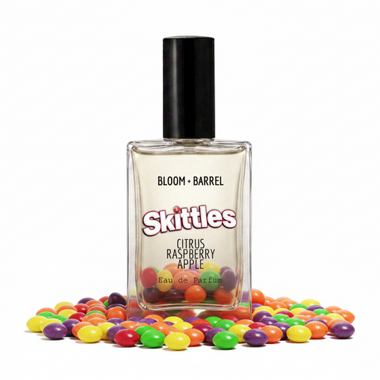 A rectangular glass perfume bottle labeled 'BLOOM • BARREL' and 'Skittles Citrus Raspberry Apple Eau de Parfum' with a black cap, shown on a white background with colorful candy-like spheres behind it.